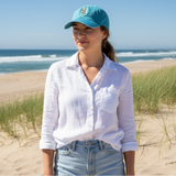 Woman wearing a blue cap and white shirt standing on a sandy beach with grass and ocean in the background.
