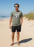 Man standing on a sandy beach wearing a green t-shirt and black shorts, holding a water bottle.