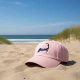 Pink cap with Cape Cod logo in navy on a sandy beach with ocean and sky in the backgroundi