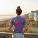 Person wearing a purple t-shirt with a Cape Cod logo, standing on a beach at sunset.